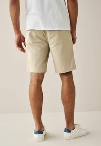 Pantaloni corti beige realizzati in tessuto, con fondo orlato. Indossati con sneakers bianche in evidenza con una linguetta blu sul tallone. Visti da dietro.