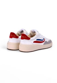 Hvite sneakers med en grå semsket skinnpanel, røde og blå detaljer, og en tykk kremfarget såle. "MADE IN PORTUGAL" preget på den røde hælen.