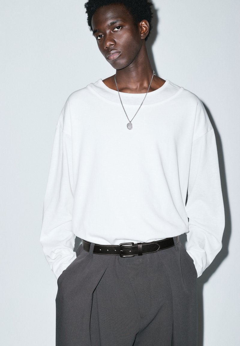 Jeune homme portant une chemise blanche ample à manches longues, un pantalon plissé gris, une ceinture noire et un pendentif, debout les mains dans les poches.