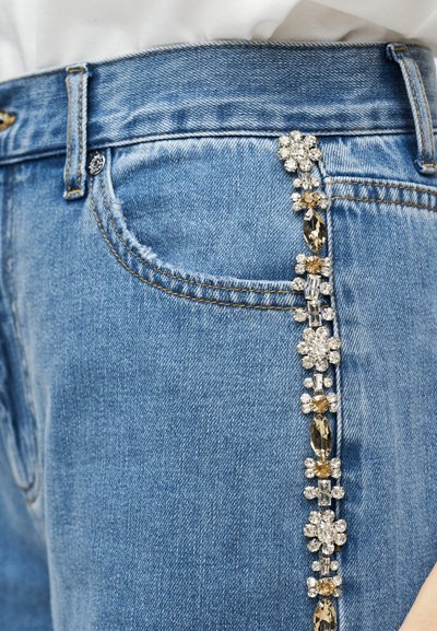 Μπλε τζιν με κάθετη σειρά διακοσμητικών διάφανων και κεχριμπαρένιων στρας κατά μήκος της άκρης της τσέπης, φορεμένα με λευκό τοπ.