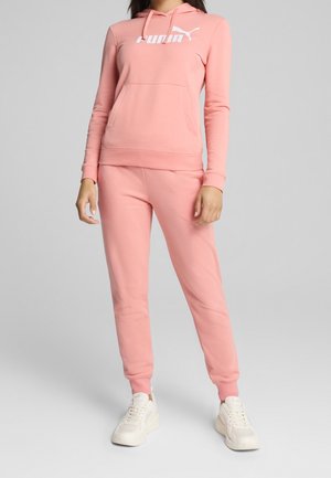 Femme portant un sweat à capuche rose Puma et un pantalon de jogging assorti avec des baskets blanches, debout contre un fond gris clair uni.