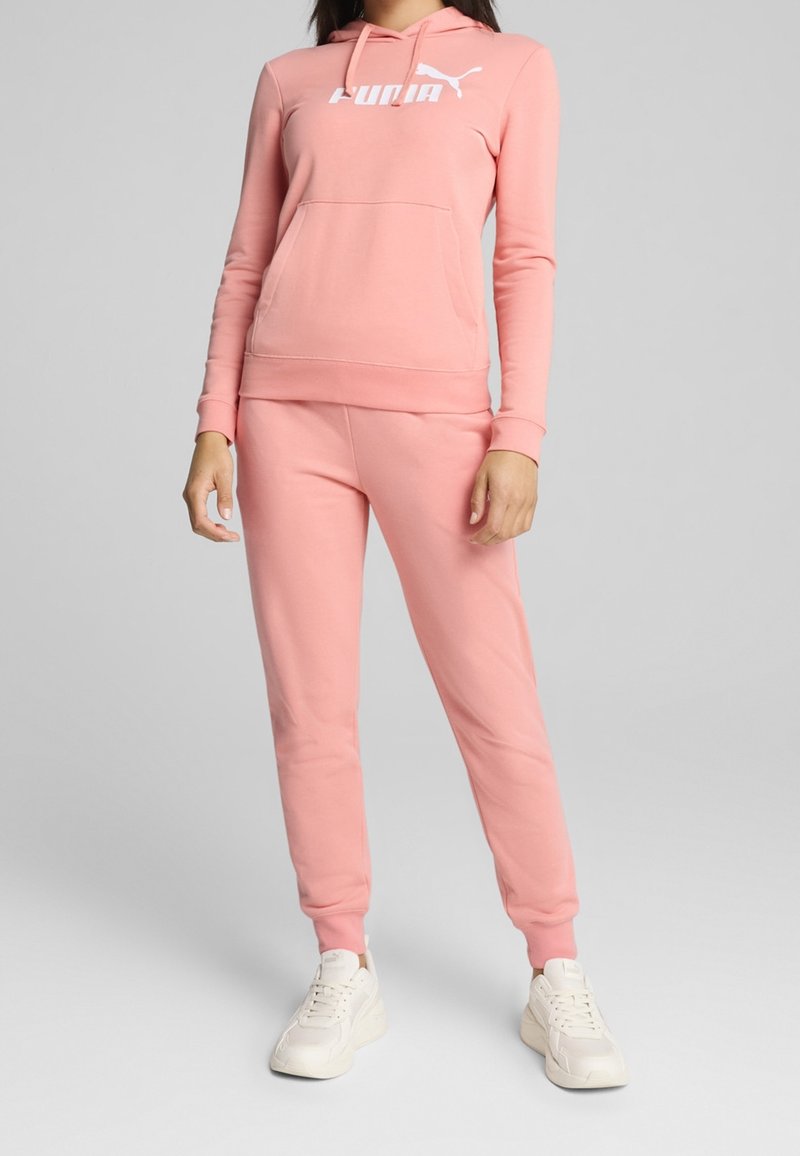Femme portant un sweat à capuche rose Puma et un pantalon de jogging assorti avec des baskets blanches, debout contre un fond gris clair uni.