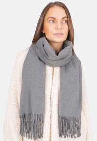 Femme portant un épais pull en maille crème et une grande écharpe grise à franges enroulée autour du cou, regardant légèrement vers la droite.