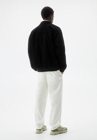 Giacca in velluto nero con orlo elastico, abbinata a pantaloni cargo bianchi a vestibilità rilassata e sneakers con dettagli verdi. Sfondo neutro.