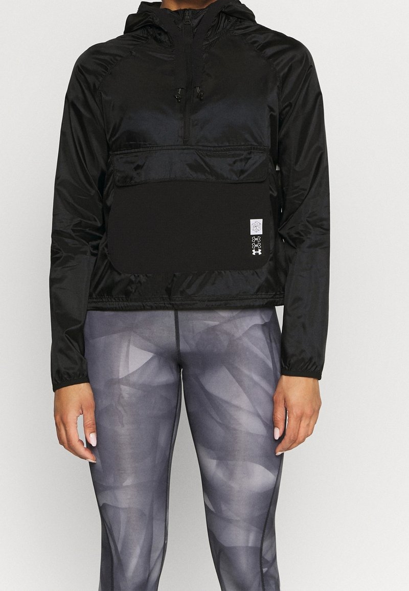 Czarna lekka kurtka z kapturem, z przednią kieszenią, elastycznymi mankietami i naszywką z logo. Połączona z szarymi legginsami w geometryczny wzór.