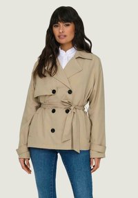 Trench coat beige con vita con cintura, bottoni neri doppi, ampie revers e maniche lunghe. Abbinato con una camicia bianca con colletto.