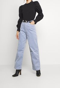 Persona che indossa una blusa nera a maniche lunghe e vaporose, pantaloni a vita alta larghi di colore azzurro chiaro con cintura, e stivaletti neri con tacco.