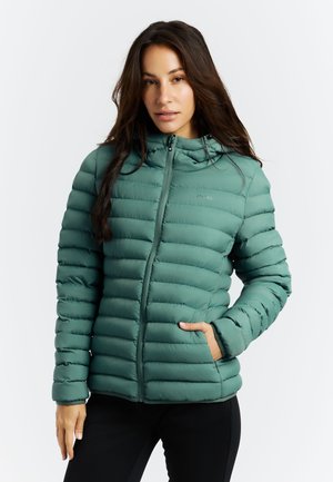 Junge Frau trägt eine grüne gesteppte Kapuzenjacke mit den Händen in den Taschen und steht vor einem schlichten weißen Hintergrund.