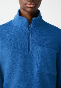 Fleece pullover i lys blå, med en halv lynlås krave, en frontlomme og tekstureret stof med et blødt, fluffy udseende.