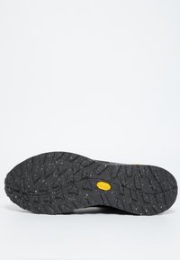 Schwarze Gummisohle mit strukturiertem Profil, das tiefe Rillen und kleine Spritzer zeigt. In der Mitte befindet sich ein gelbes Markenemblem.