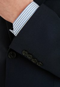 Morska marynarka z mankietem zdobionym pięcioma czarnymi guzikami; pod spodem widoczny mankiet jasnoniebieskiej koszuli w białe paski. Szczegół tkaniny o fakturze.