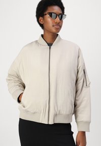 Lys beige bomberjakke laget av mykt stoff, med ribbede mansjetter, glidelås foran og en sidelomme med glidelås.