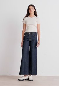 Hvit kortermet topp kombinert med høyt midjebånd og vide mørkeblå denimbukser. Modellen har på seg hvite sko. Enkel bakgrunn.