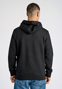 Svart huvtröja med mjuk textur, känguruficka och ribbade ärmar. Buren med ljusblå jeans, sett från baksidan.
