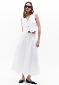 Weißes, kurzes Top mit goldenen Knöpfen und tiefem V-Ausschnitt, kombiniert mit einem fließenden weißen Maxirock und schwarzen Ballerinas. Glattes, leichtes Material.
