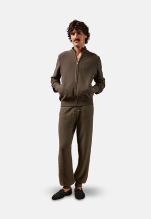 Ensemble de survêtement en maille couleur vert olive comprenant une veste zippée et un pantalon à cordon avec poignets côtelés. Tissu texturé avec un motif subtil. Chaussures noires à enfiler.