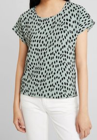 Světle zelený krátký rukáv top s kulatým výstřihem a černým oválným tečkovaným vzorem, vyrobený z hladkého materiálu. V kombinaci s bílými kalhotami.