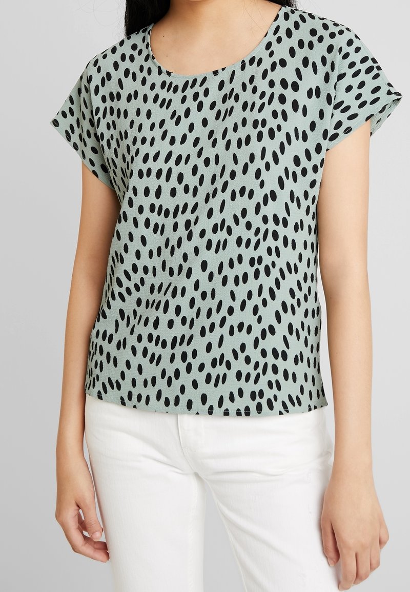 Světle zelený krátký rukáv top s kulatým výstřihem a černým oválným tečkovaným vzorem, vyrobený z hladkého materiálu. V kombinaci s bílými kalhotami.