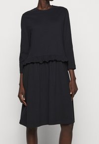 Svart långärmad klänning med volangsläpp, tillverkad av mjukt tyg. A-linjeform med rund halsringning och subtila sömdetaljer.