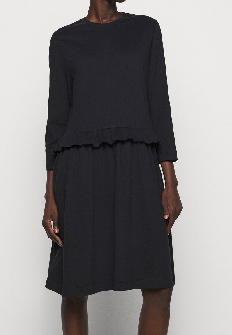 Svart långärmad klänning med volangsläpp, tillverkad av mjukt tyg. A-linjeform med rund halsringning och subtila sömdetaljer.