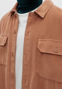Detailní záběr na osobu, která má na sobě hnědou kordovou košili na knoflíky přes bílou košili a tmavozelený rolák.
