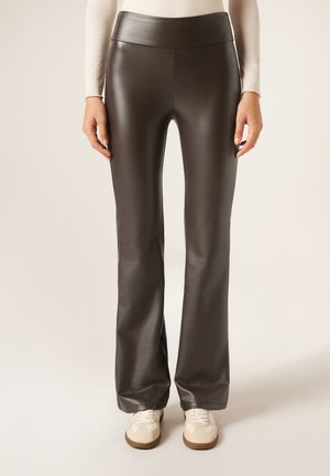 Person iført mørkebrune bukser i kunstlæder med vidde forneden og beige sneakers, stående mod en ensartet lys baggrund.