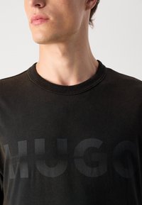 Juodas medvilnės marškinėlis su apvaliu kaklu, trumpomis rankovėmis ir dideliu, nykstančiu "HUGO" logotipu, atspauduotu ant krūtinės.