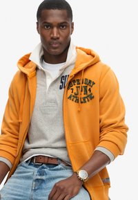 Felpa arancione con zip e dettagli grigi, indossata sopra un polo grigio chiaro, abbinata a jeans azzurri e un orologio dorato.