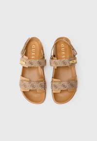 Coppia di sandali color cuoio con cinturini decorati, logo dorato "GUESS" sui cinturini alla caviglia e suole imbottite, mostrati su uno sfondo bianco.