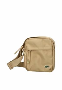 Borsa crossbody beige con tracolla regolabile, tasca frontale con zip e piccolo logo di coccodrillo verde nell'angolo in basso a destra.