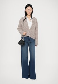 Cardigan grigio fluffy con grandi bottoni, abbinato a jeans a gamba larga blu. Porta una piccola borsa a pochette nera. Design minimalista.