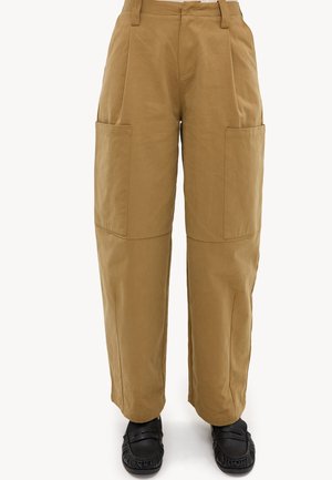 Pantalones marrones de corte recto con grandes bolsillos delanteros, usados con zapatos negros sin cordones sobre un fondo blanco.
