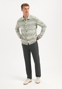 Licht gestructureerd overhemd met groene, witte en blauwe strepen. Gecombineerd met donkergrijze broek en beige sneakers op een effen achtergrond.