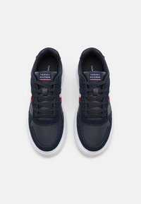 Pár námořnických modrých tenisek Tommy Hilfiger s bílou podrážkou, červenými a bílými bočními pruhy a černými tkaničkami, pohled zhora na bílém pozadí.