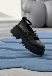 Scarpe platform in pelle nera con tre fibbie, suola spessa dentellata e punta arrotondata. Mostrate su un tappeto blu testurizzato.