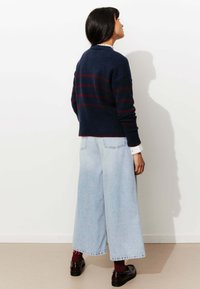 Pull-over marine avec des rayures rouges, design court, poignets côtelés, associé à un jean taille haute bleu clair et des chaussures noires, vu de derrière.