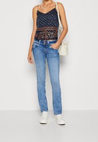 Navy floral print camisole with spaghetti straps, paired with light blue straight-leg jeans and white sneakers, holding a beige handbag.