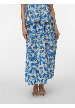 Kvinde iført beige nederdel og top med kraftige blå blomsterprint, kombineret med beige sandaler med stropper, stående foran en hvid baggrund.