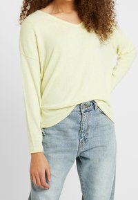 Jasnożółta bluzka z długim rękawem, dekoltem w serek, luźnym krojem i zakrzywionym dołem. W zestawieniu z jasnoniebieskimi dżinsami z wysokim stanem. Miękka faktura.