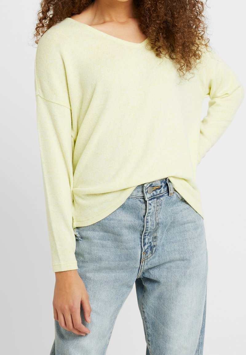 Jasnożółta bluzka z długim rękawem, dekoltem w serek, luźnym krojem i zakrzywionym dołem. W zestawieniu z jasnoniebieskimi dżinsami z wysokim stanem. Miękka faktura.
