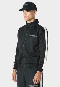 Czarna kurtka dresowa na zamek z białymi paskami po bokach, wyposażona w ściągacze przy mankietach i elastyczny dół. Połączona z pasującymi czarnymi spodniami, obie rzeczy mają detale z logo.