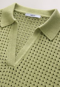 Polo in maglia traforata verde chiaro, con scollo a V, colletto e motivo lavorato a trama con piccoli fori su tutto il tessuto.