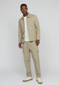 Mann in beiger Jacke und Hose mit weißen Sneakers, der vor einem grauen Hintergrund steht.