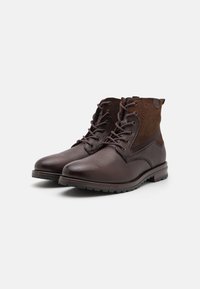 Botas de tobillo de cuero marrón con un acabado suave, acentos de ante marrón contrastante, punta redonda y suela de goma resistente. Diseño con cordones.