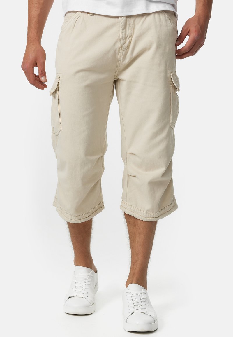 Shorts cargo beiges en tissu coton, avec poches latérales, longueur au genou et détails cousus. Portés avec des baskets blanches.
