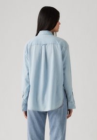 Camicia di jeans azzurra chiara con maniche lunghe, polsini con bottoni, cucitura del yoke posteriore, orlo curvo più lungo e vestibilità rilassata. Abbinata a jeans azzurri chiari.