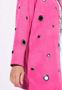 Růžová semišová bunda s uvolněným střihem, která má velké černé kroužky uspořádané asymetricky na rukávech a těle.