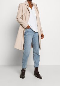 Beige lang frakke med knapdetaljer, hvid V-udskæring top, blå lige ben jeans og mørkebrun ankelstøvler med tekstureret overflade.