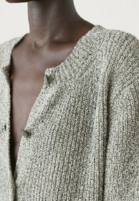 Cardigan beige con trama, caratterizzato da un profondo scollo a V, collo a coste e due bottoni scuri vicino al colletto. Materiale morbido con vestibilità ampia.