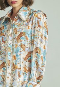 Blusa multicolore con un motivo floreale blu, marrone e crema. Include bottoni simili a perle e rifiniture decorative lungo il colletto e i bottoni.